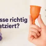 Eine Hand hält eine orange Menstruationstasse vor hellem Hintergrund mit lila Text "¿Copa bien colocada?".