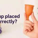 An orange menstrual cup is held by a hand, alongside the question "¿Copa bien colocada?" and stacked towels.