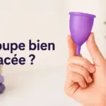 Une main tient une coupe menstruelle violette devant un texte questionnant son placement, et des linges pliés.