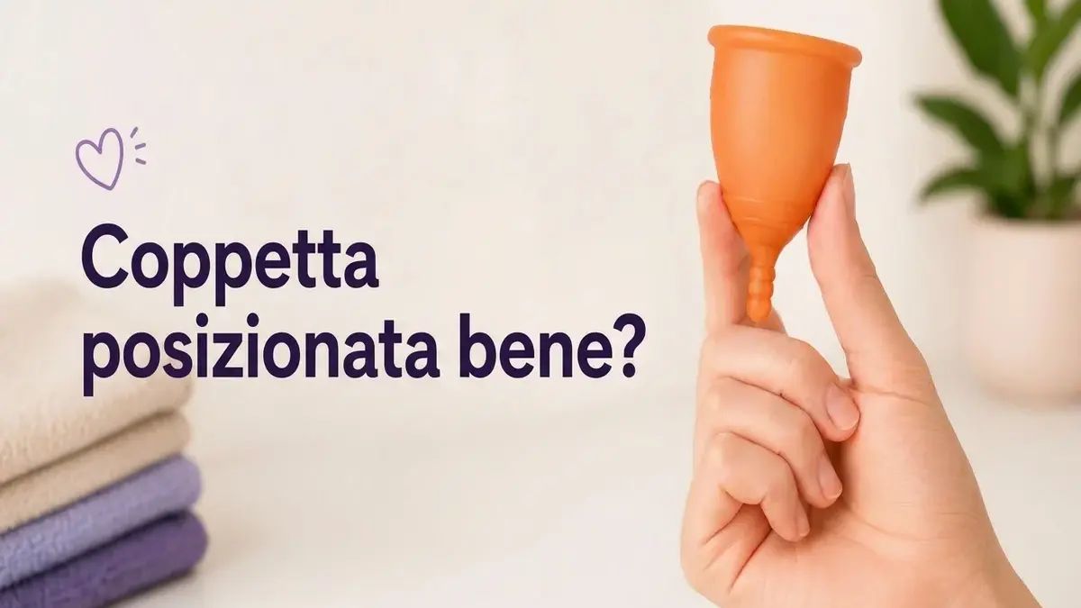 Mano che mostra una coppetta mestruale arancione con il testo "¿Copa bien colocada?" e asciugamani sullo sfondo.