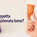 Mano che mostra una coppetta mestruale arancione con il testo "¿Copa bien colocada?" e asciugamani sullo sfondo.