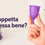 Mano che tiene una coppetta mestruale viola, con la domanda "¿Copa bien colocada?" e asciugamani sullo sfondo.